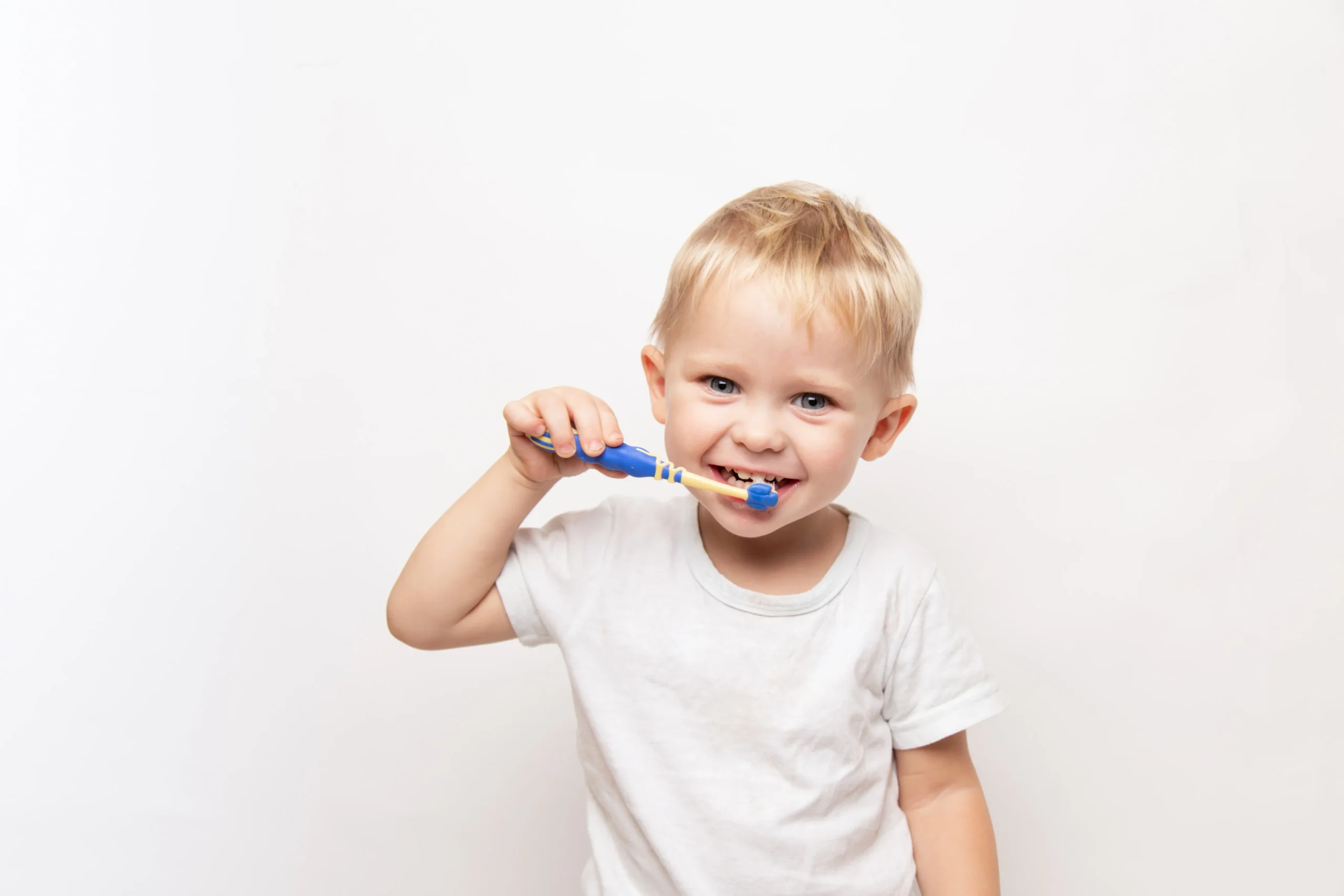 little cute caucasian blond blue-eyed boy in a white t-shirt brushes his teeth on a white background.