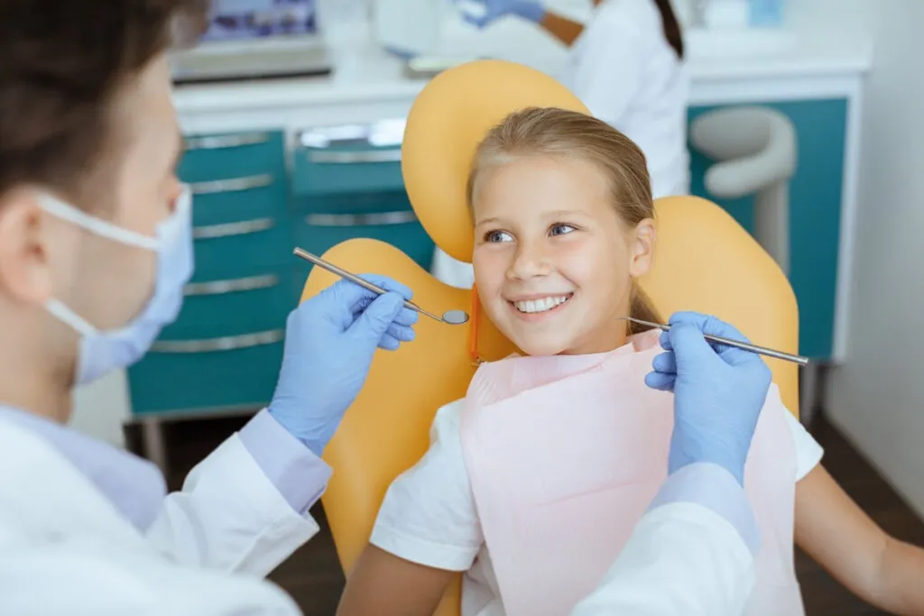 Fearless visit to doctor and dental treatment for child. Smiling girl in medical chair looks at dentist man in white coat, protective mask and rubber gloves with tools in his hands, and not afraid