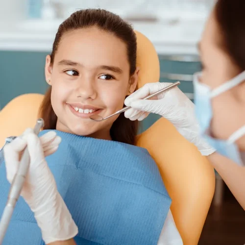 Dental care prevention and treatment procedure. Millennial female doctor in protective mask and gloves holds medical instrument and checks oral cavity of little smiling girl in modern clinic interior